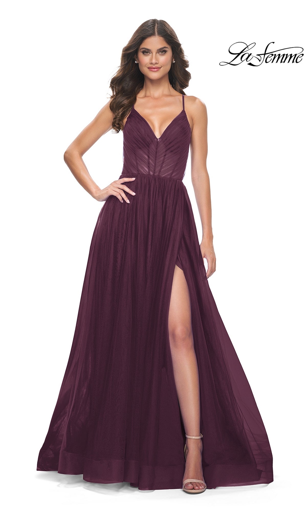 A woman in the La Femme Sheer-Waist Long Prom Ball Gown 31457, a sleeveless deep burgundy dress with thin straps, a fitted bodice, and a slit, stands against a plain background—a stunning twist on the classic ball gown.